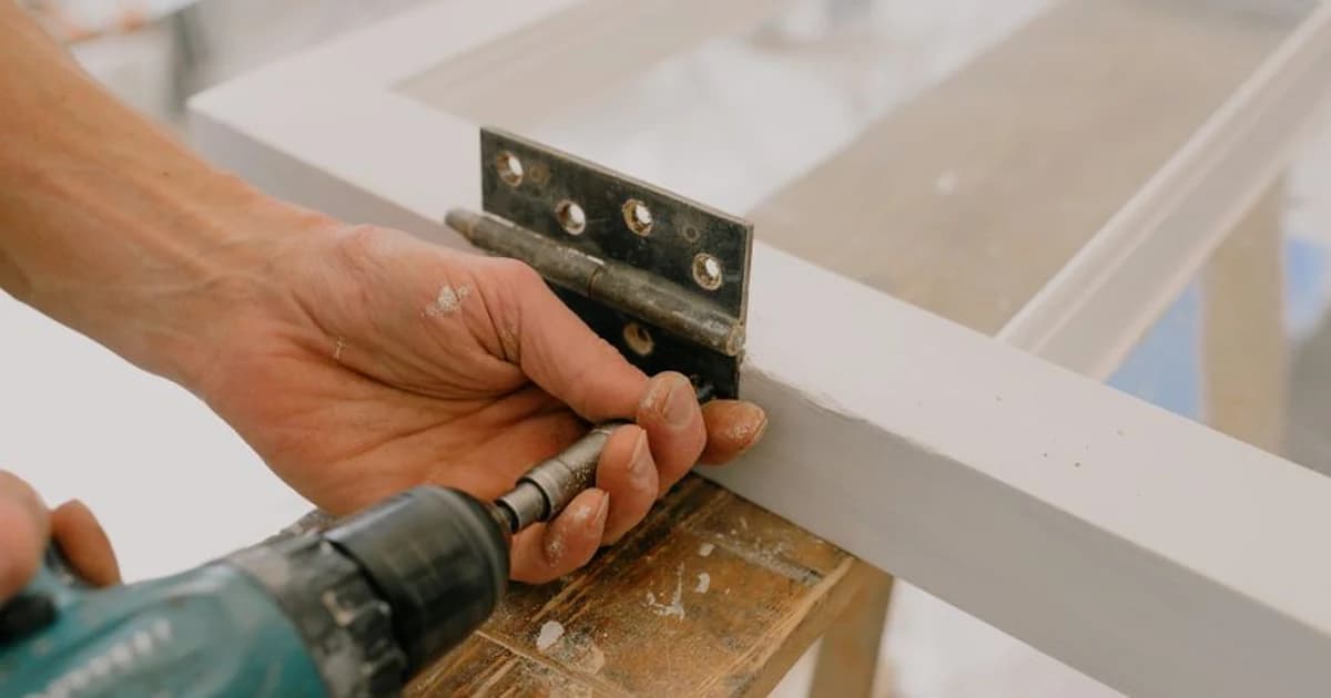 A hand driving a screw into a door hinge with a cordless drill