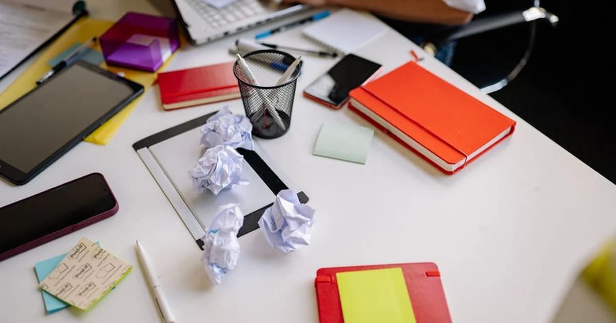 A desk covered with crumpled paper, sticky notes and notebooks beside a laptop