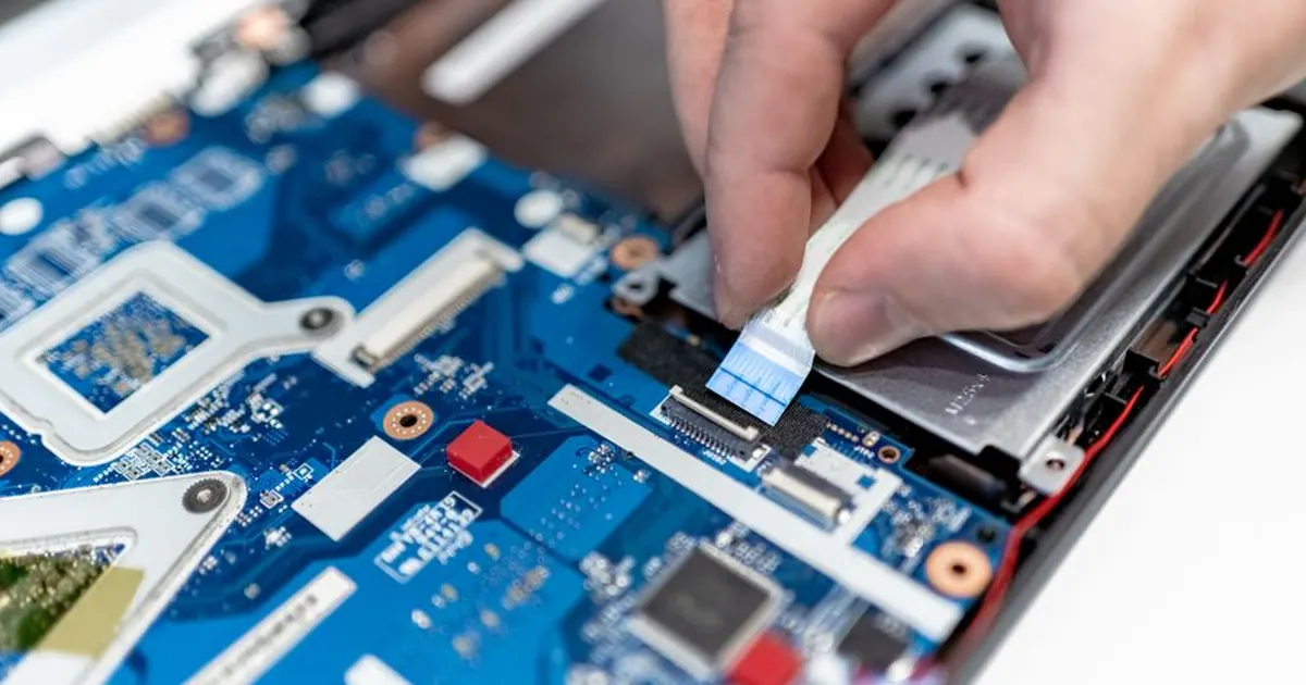 Hands reconnecting a ribbon cable on the exposed circuit board of an opened laptop