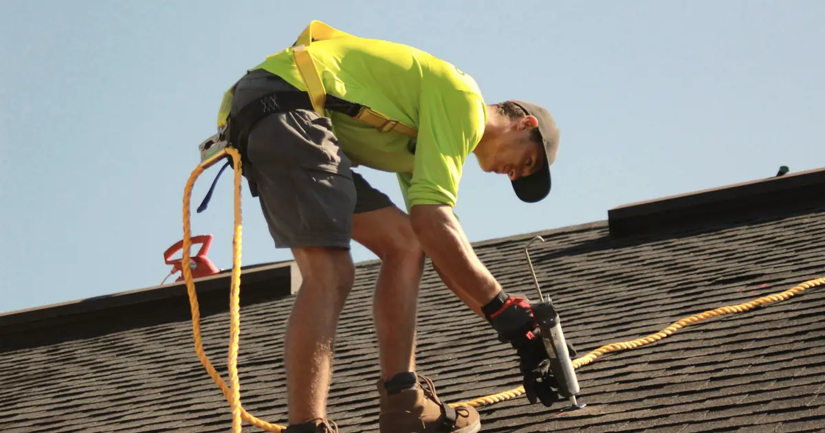 Roofer in a safety harness applying sealant to an asphalt shingle roof under a clear sky