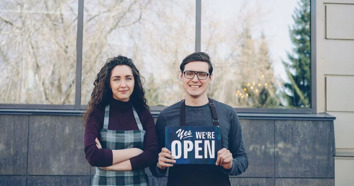 Two small business owners standing outside their storefront holding a Yes We're Open sign